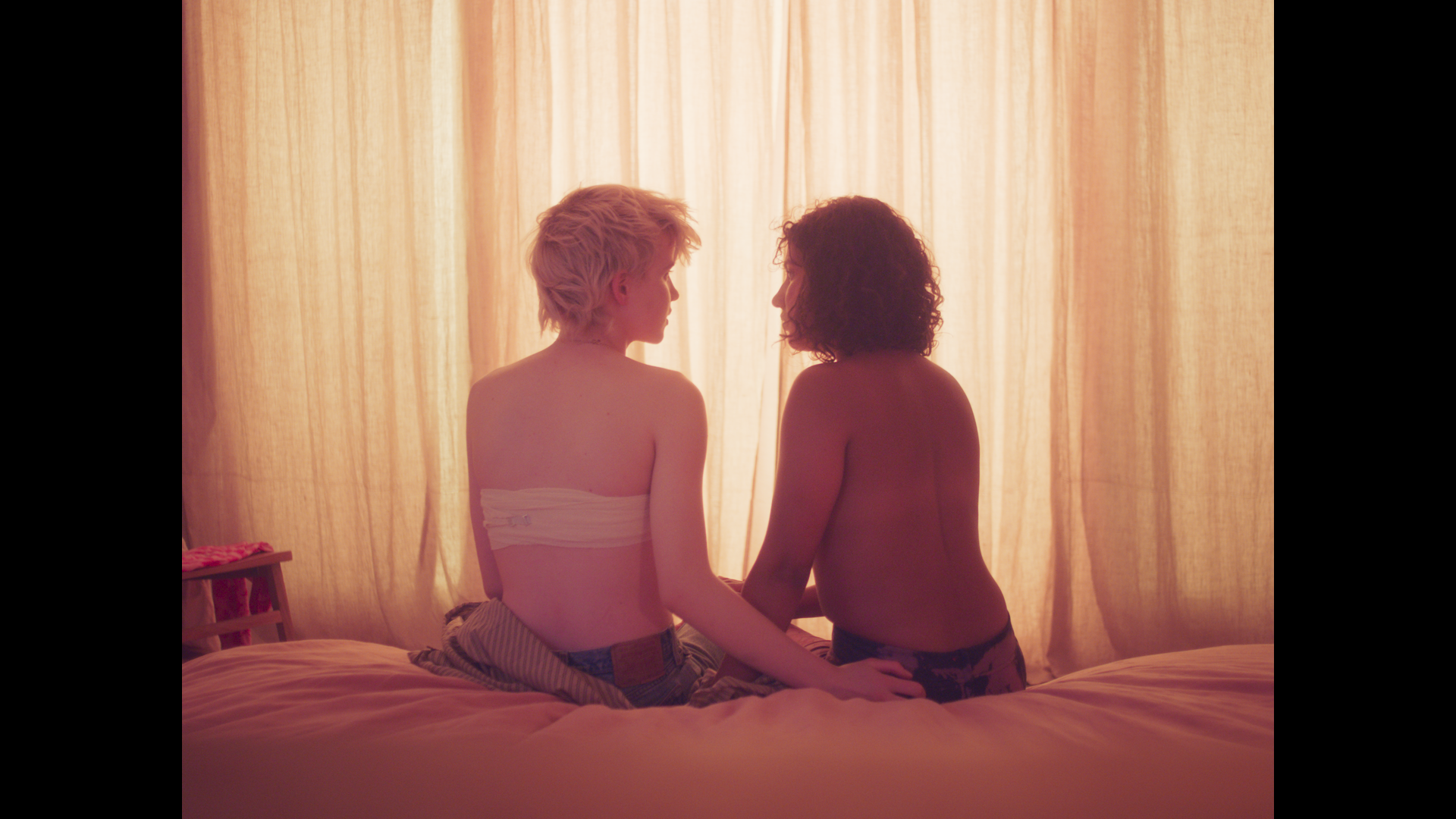 Zwei junge Menschen sitzen ohne Oberteil von der Kamera abgewandt auf einem Bett. Eine Person hat Verbände im Brustbereich.
