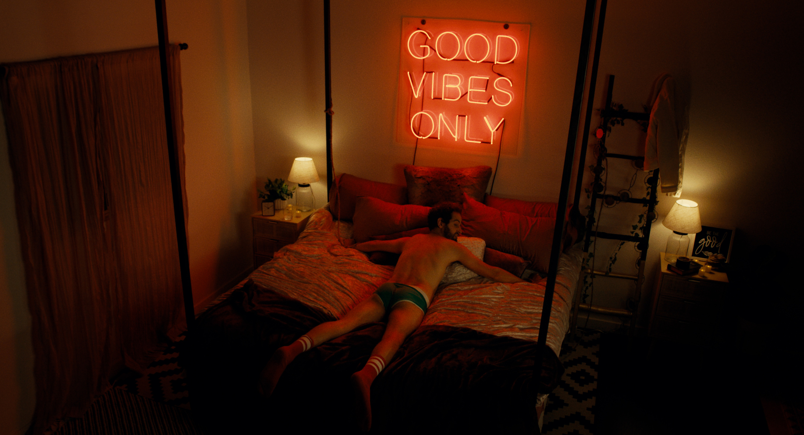 Mann liegt in Unterwäsche auf dem Bett. Über sich Neon-Buchstaben: Good vibes only.